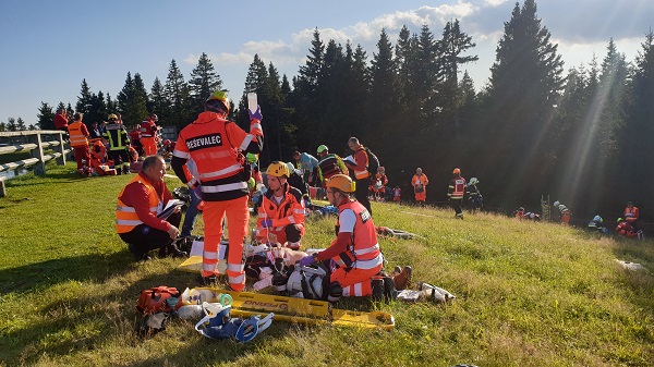 Slika ekipe NMP Ajdovščina, sestavljena iz treh članov, poleg še ocenjevalec, ki ocenjuje izvajanje enega izmed scenarijev. Dogodek poteka na travnatem pobočju, v odzadju tudi ekipe drugih tekmovalcev ter iglasti godz