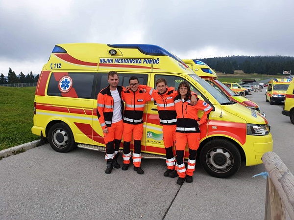 Slika ekipe NMP sestavljena iz štirih članov, na Tekmovanju ekip NMP Rogla 2019, ki stoji pred reševalnim vozilom, v odzadju kolona reševalnih vozil